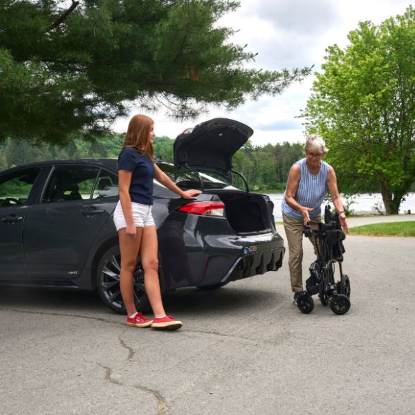 A woman in a navy blue shirt and white shorts stands by the open trunk of a dark gray sedan, while an older woman pushes a folded Golden Technologies Cricket LT Carbon Lightweight Power Wheelchair beside the car on a paved outdoor area with trees in the background.
