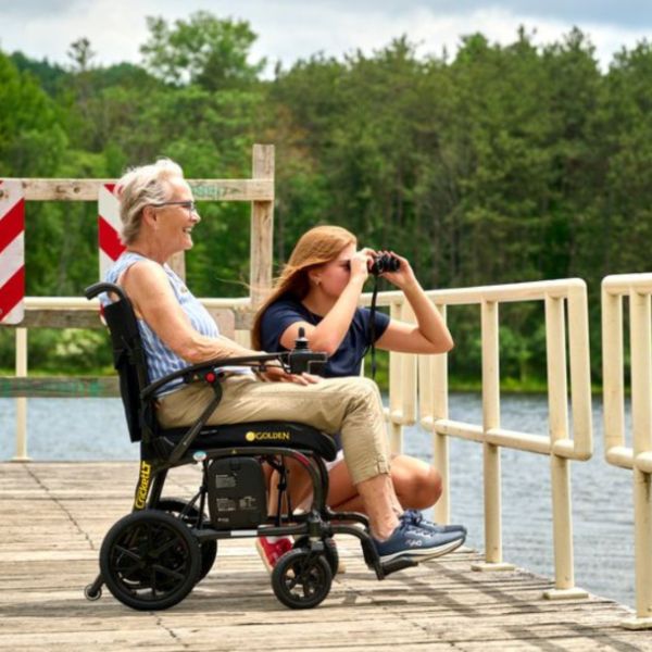 A woman in a Golden Technologies Cricket LT Carbon Lightweight Power Wheelchair sits on a wooden dock by the water, talking to another woman who is holding binoculars. Trees and water are visible in the background.