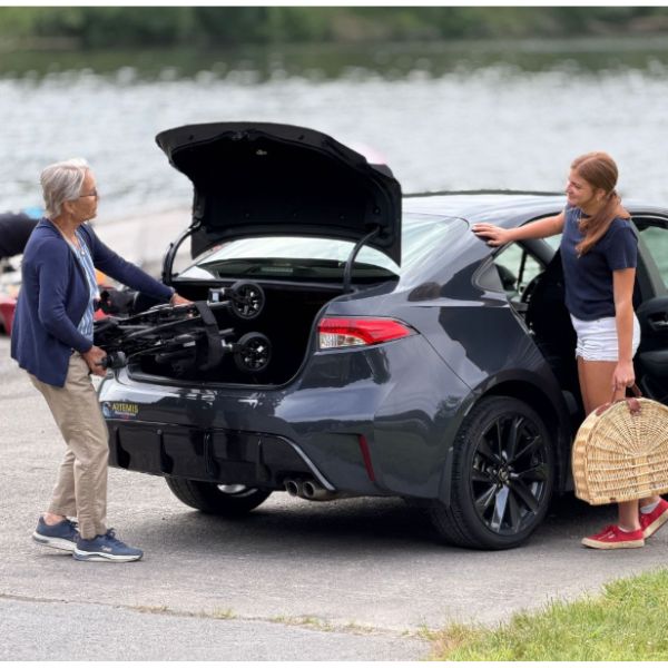 A woman in a navy cardigan and beige pants loads the Cricket LT Carbon wheelchair into the open trunk of a dark gray sedan, while a woman in a navy t-shirt and white shorts stands by the open car door holding a woven basket. The scene is set outdoors next to a body of water.